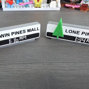 May include: Two rectangular signs, one reading "TWIN PINES MALL" and the other "LONE PINE MALL". Each sign has a green tree and a digital clock displaying the time. The signs are gray with white and black details.