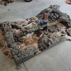 Könnte beinhalten: Ein detailliertes Diorama eines felsigen Geländes mit einem kleinen blauen Spielzeug-LKW. Die Landschaft zeigt graue Felsformationen, eine Holzbrücke und ein beigefarbenes Flussbett. Das Diorama ist für Modellautos und andere kleine Spielzeuge konzipiert.
