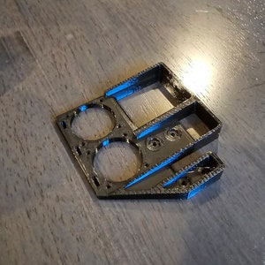 May include: Black 3D-printed plastic component with geometric shapes. Features two circular openings, a rectangular section, and angled slots. The object is designed with a textured surface and appears to be a part of a larger assembly.
