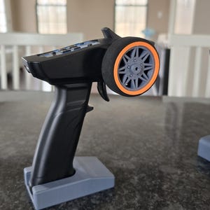 May include: A black remote control with an orange-rimmed tyre on the top. The remote control is sitting on a grey stand.