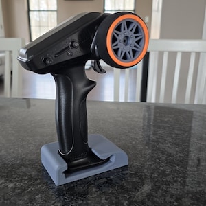 May include: Black remote control with an orange and grey wheel, resting on a grey stand. The remote has a trigger and several buttons. The wheel has a star-like design. The stand is rectangular with a curved opening.
