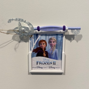 May include: A white plastic frame with a picture of the characters from the movie Frozen II. The frame is attached to a purple and white plastic hanger with a sparkly blue Disney logo. The text on the frame reads "Disney Frozen II shopDisney.com | Disney store".