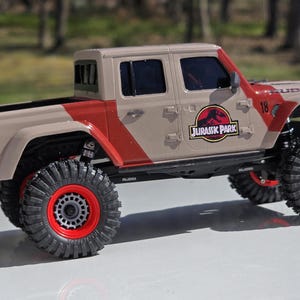 Può includere: Un camion giocattolo beige e rosso con grandi pneumatici neri e cerchi rossi. Il camion ha la scritta "JURASSIC PARK" e il numero "18" sul lato. È un modello dettagliato di un pick-up.