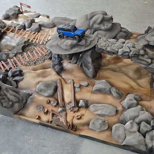 Puede incluir: Diorama detallado de un circuito todoterreno en miniatura con un camión de juguete azul. El circuito presenta formaciones rocosas grises, un puente de madera y un terreno arenoso. Un pequeño letrero marca la línea de meta.