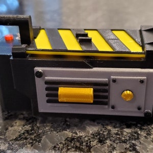 May include: A black and yellow plastic toy proton pack with a handle, a red button, and a gray panel with a yellow knob and a black grill.