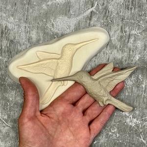 May include: A hummingbird sculpture in a light gray color, next to a matching mold. The bird has detailed wings and a long beak. The mold is off-white and shaped to the bird's form. The background is a textured gray surface.