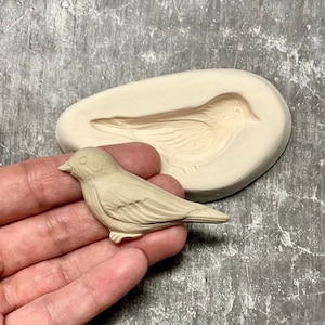 Peut inclure: Une figurine d'oiseau en céramique beige et un moule en silicone du même oiseau. Le moule est de forme ovale et a une surface texturée.