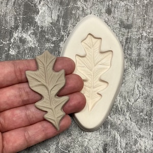 May include: A tan-colored oak leaf-shaped mold and a cast leaf. The mold is white and has a leaf impression. The cast leaf is a lighter tan color. The items are likely for crafting or decorative purposes.