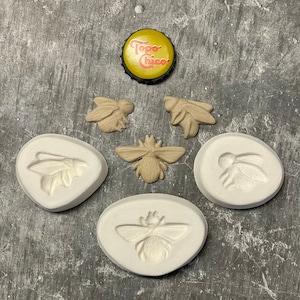 May include: An overhead shot features a yellow and black Topo Chico bottle cap, along with several bee-themed craft molds and bee-shaped embellishments. The molds are white, and the bees are tan.