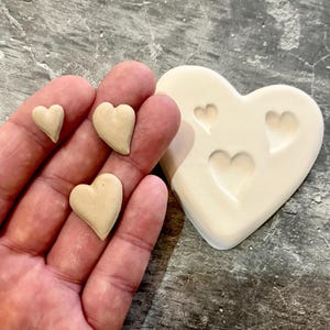 May include: Three small heart-shaped clay pieces next to a heart-shaped mould with three heart-shaped impressions. The clay pieces are a light beige colour and the mould is a slightly darker beige.