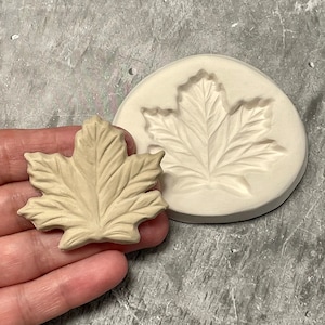 Peut inclure: Un moule en forme de feuille d'érable beige et une feuille moulée. La feuille moulée est tenue dans une main. Le moule est rond et présente une empreinte de feuille détaillée. La feuille et le moule sont sur une surface grise. La feuille mesure environ 7.6 cm de long.