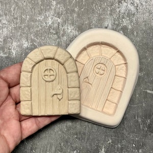 May include: A beige, miniature fairy door with a round top and a small, round window. The door has a handle and is next to a matching mold. The door is made of a light-colored material.