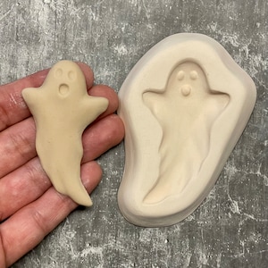 May include: A beige ghost-shaped ceramic mold and a finished ghost figurine. The ghost has a surprised expression with wide eyes and an open mouth. The mold is a light beige color, and the finished ghost is held in a hand.