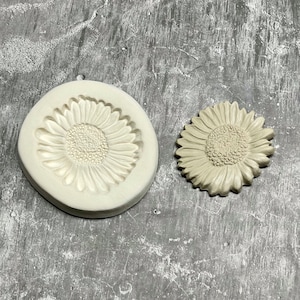 Op de afbeelding: Een witte siliconen mal en een witte keramische zonnebloem, beide met een reliëfdesign. De mal is een negatief beeld van de zonnebloem.