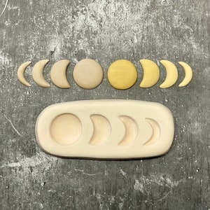 Pode incluir: Um conjunto de seis moldes de resina de fase lunar, incluindo formas de lua cheia, lua crescente e meia lua. Os moldes são feitos de um material de silicone bege claro e estão dispostos em uma fila sobre um fundo cinza.