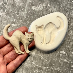 Peut inclure: Une figurine de chat beige clair avec le dos arqué et une expression souriante. À côté, un moule assorti. Le chat est debout sur ses quatre pattes, la queue courbée vers le haut. La figurine mesure environ 7.6 cm de long.
