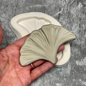 Puede incluir: Un molde de silicona gris claro en forma de hoja de ginkgo. El molde se muestra con una fundición de hoja de ginkgo terminada.