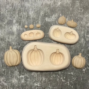 May include: Beige pumpkin molds and individual pumpkins are arranged on a gray surface. The molds vary in size, each with pumpkin-shaped cavities. Individual pumpkins of various sizes are also present, suggesting a fall or harvest theme.