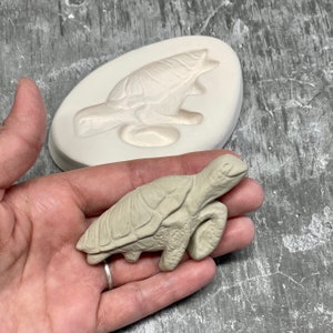 May include: A white silicone mold of a turtle, with a small, gray ceramic turtle next to it.