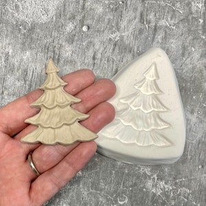 Könnte beinhalten: Ein beigefarbener Weihnachtsbaumschmuck neben einer weißen Form. Der Schmuck, gehalten in einer Hand, weist ein detailliertes, geschichtetes Design auf. Die Form spiegelt die Form des Baumes wider, was auf ein Bastel- oder DIY-Thema hindeutet.