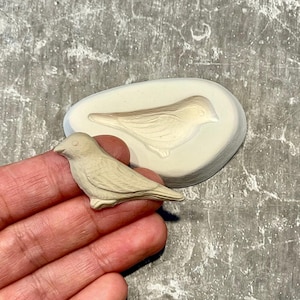 May include: A small, beige bird figurine next to a matching bird-shaped mold. The bird is detailed with visible wings and feathers. The mold is oval-shaped, with a bird impression. The objects are on a textured gray surface.