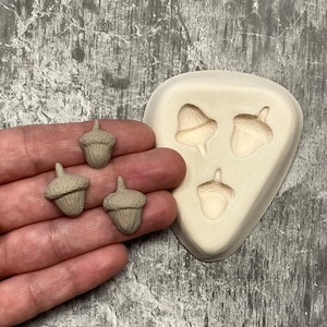 Peut inclure: Gros plan sur trois petits objets en forme de glands de couleur beige tenus dans une main. Un moule blanc avec trois formes de glands est en arrière-plan. Les glands ont des chapeaux détaillés et des corps texturés.