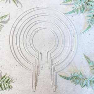 May include: A white concrete surface with a design of concentric circles and a vertical line in the center. The design is reminiscent of a mandala or a zen garden. Green leafy plants are placed around the design.