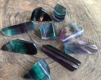 May include: A collection of polished fluorite stones in various shapes and sizes. The stones display a mix of green, purple, and clear colors. The stones are arranged on a natural wood surface.