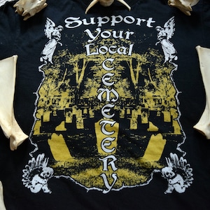 May include: Black t-shirt with a graphic print of a cemetery with the text "Support Your Local Cemetery" in white lettering. The graphic is surrounded by images of angels.