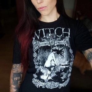 Peut inclure: T-shirt noir avec un motif blanc d'une femme chevauchant un balai. Le motif est encadr&eacute; d'une bordure d&eacute;corative et le mot "WITCH" est imprim&eacute; au-dessus du motif.