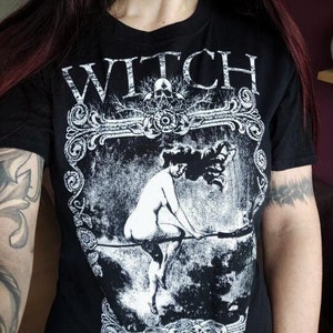Peut inclure: T-shirt noir avec un motif blanc d'une femme sur un balai. Le motif est entour&eacute; d'un cadre d&eacute;coratif et le mot "WITCH" est imprim&eacute; au-dessus.
