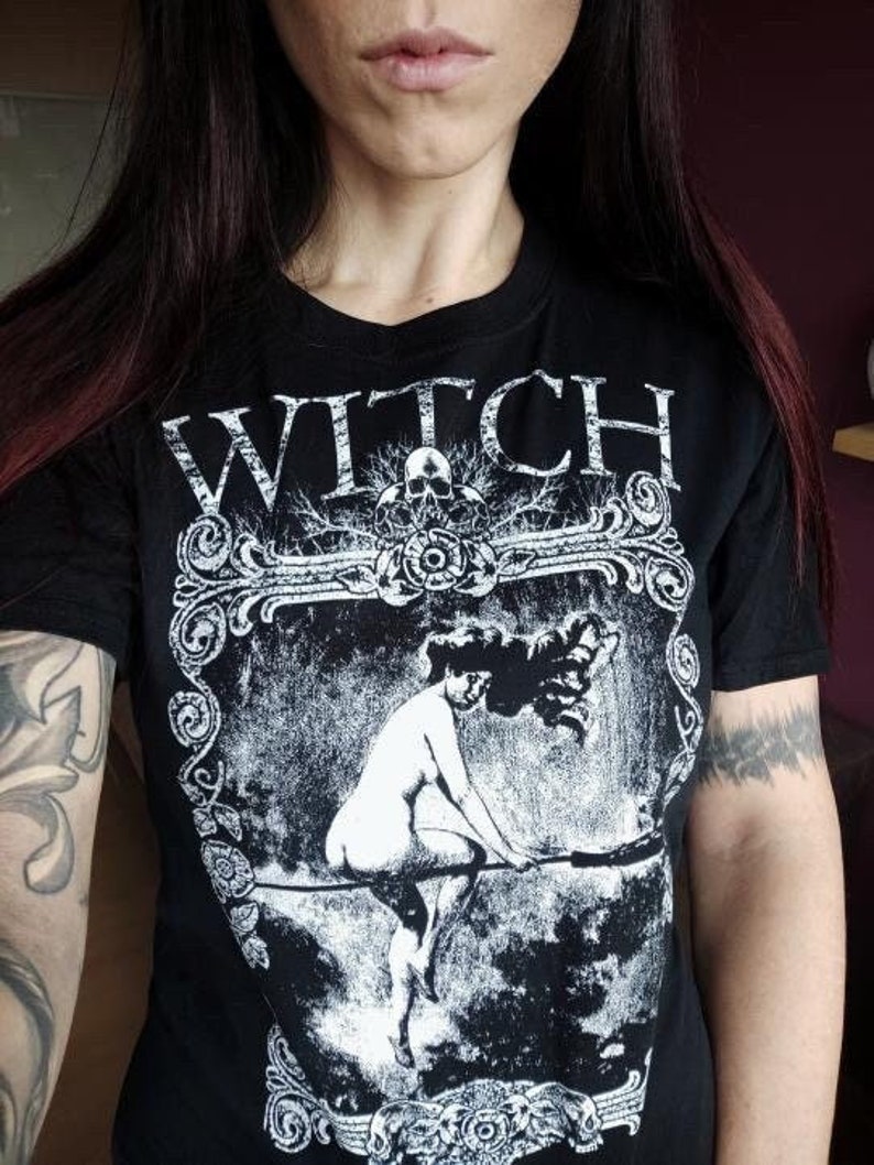 Peut inclure: T-shirt noir avec un motif blanc d'une femme sur un balai. Le motif est entour&eacute; d'un cadre d&eacute;coratif et le mot "WITCH" est imprim&eacute; au-dessus.