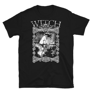 Peut inclure: T-shirt noir avec un graphique blanc d'une sorci&egrave;re chevauchant un balai dans un cadre d&eacute;coratif. Le texte "WITCH" est au-dessus du cadre.