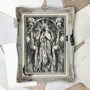 May include: A framed black and white print depicting a woman in a long dress standing in a forest, flanked by two figures. The figures are partially obscured by the trees and have a haunting, ethereal quality. The print is in a silver frame with ornate details.