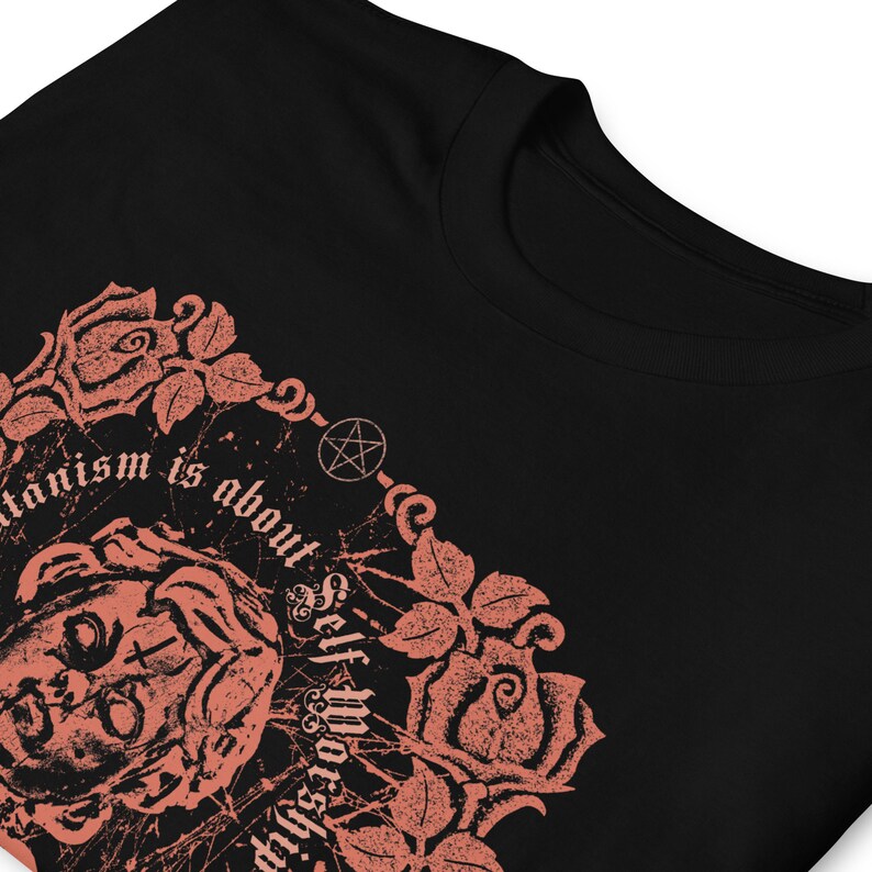 May include: Black t-shirt with a pink graphic design featuring a stylized face with a pentagram and roses. The text reads "Satanism is about self worship".