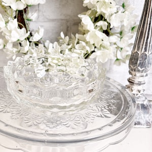 Puede incluir: Un cuenco de vidrio transparente con un borde festoneado se encuentra sobre un soporte para pasteles de vidrio transparente con un patrón decorativo. El cuenco está vacío y parece estar hecho de vidrio grueso.