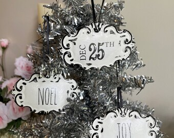 May include: Three white metal Christmas ornaments with black lettering. The ornaments are shaped like a banner and have a distressed look. The ornaments say "DEC 25TH", "NOEL", and "JOY".