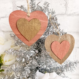 Peut inclure: Deux décorations en bois en forme de cœur avec un design superposé. Le plus grand cœur est de couleur rose clair et le plus petit cœur est de couleur marron. Les décorations sont suspendues à un arbre en tinsel argenté.