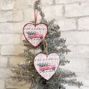May include: Two pink heart-shaped Christmas decorations featuring a red truck carrying a Christmas tree on each. The decorations are made of wood and have a vintage sheet music design.