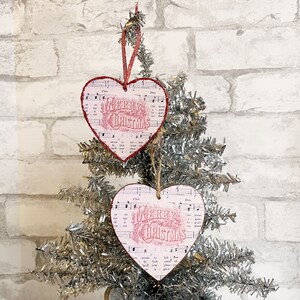 Peut inclure: Deux décorations de Noël en forme de cœur rose avec les mots "Merry Christmas" imprimés dessus. Les décorations sont suspendues à un sapin de Noël en tinsel argenté.