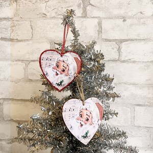 May include: Two red heart-shaped Christmas ornaments with a vintage Santa Claus image on each. The ornaments are decorated with musical notes and a holly sprig.