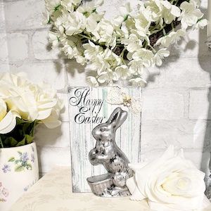 Könnte beinhalten: Ein weißes Holzschild mit der Aufschrift "Happy Easter" in schwarzen Buchstaben. Eine silberne Hasenfigur ist an dem Schild befestigt. Das Schild steht auf einem Tisch mit einer weißen Rose im Vordergrund.
