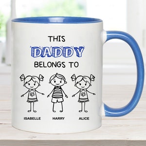 May include: White ceramic mug with a blue rim and handle. The mug has a blue inscription that reads "This Daddy Belongs To" with three stick figure drawings of children below. The children are labeled "Isabelle", "Harry", and "Alice".