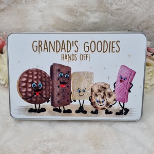 Puede incluir: Una lata de metal blanca con un borde plateado y el texto "Grandad's Goodies Hands Off!" en letras marrones. La lata presenta una ilustraci&oacute;n de dibujos animados de cinco tipos diferentes de galletas con caras, brazos y piernas.