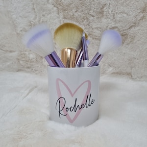 May include: White ceramic makeup brush holder with a pink heart and the name "Rochelle" written in black. The holder is filled with various makeup brushes, including a large fluffy brush, a smaller brush with a purple handle, and a few smaller brushes with white handles.