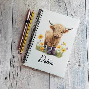 May include: A white spiral notebook featuring a brown and white Highland cow illustration wearing purple wellies. The cow is standing in a field of yellow flowers. The notebook is personalised with the name "Debbi" on the front.