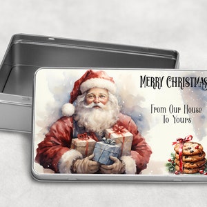 May include: A silver metal tin with a Christmas design. The tin features a watercolor illustration of Santa Claus holding gifts, with the text "Merry Christmas From Our House To Yours".
