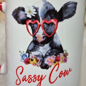 May include: White ceramic mug with a red rim. The mug features a cartoon illustration of a black and white cow wearing red heart-shaped sunglasses and a white flower. The cow is surrounded by colorful flowers. The text "Sassy Cow" is written in red cursive below the cow.