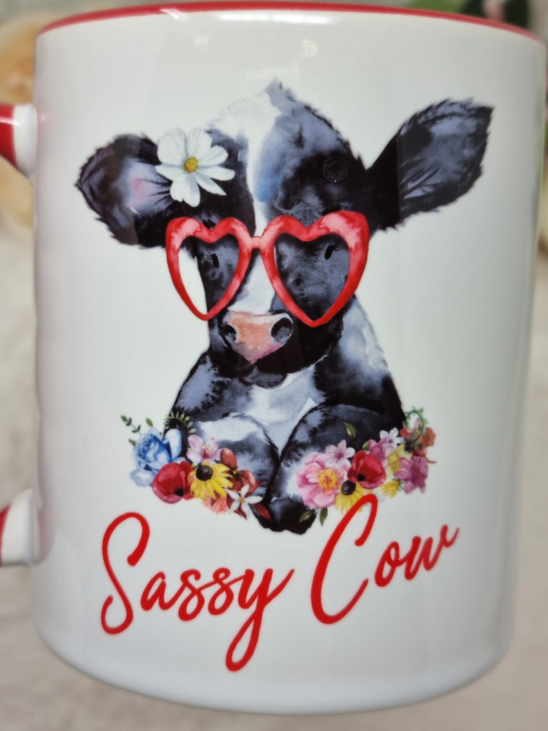 May include: White ceramic mug with a red rim. The mug features a cartoon illustration of a black and white cow wearing red heart-shaped sunglasses and a white flower. The cow is surrounded by colorful flowers. The text "Sassy Cow" is written in red cursive below the cow.
