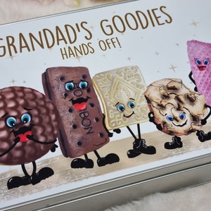 Puede incluir: Una lata de plata con fondo blanco y el texto "Grandad's Goodies Hands Off!" en letras marrones. La lata presenta personajes de dibujos animados de varias galletas con caras, brazos y piernas.